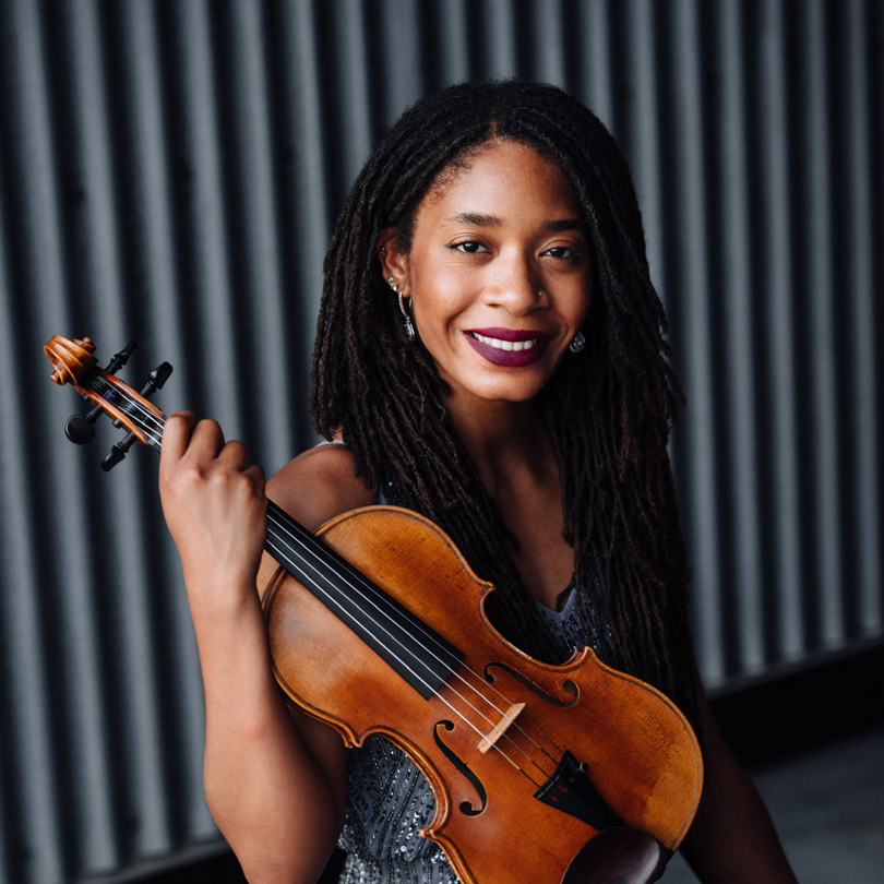 Woman with long dreadlocks smiling while holding a violin against a dark curtain backdrop.